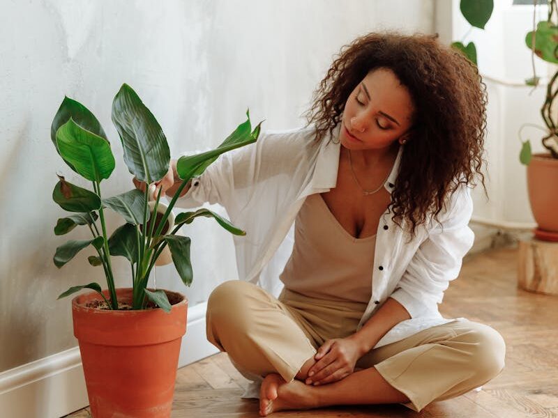 An African American woman sitting on a wooden floor, caring for a potted indoor plant in a bright, modern home.