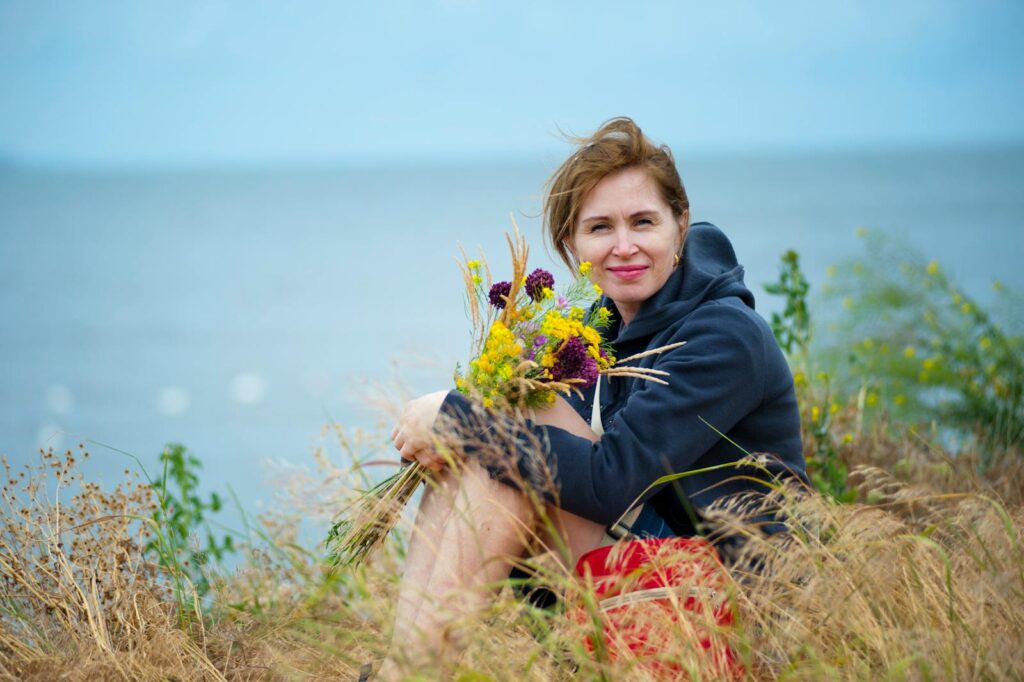Smiling woman holding wildflowers by the seaside, enjoying summer outdoors.