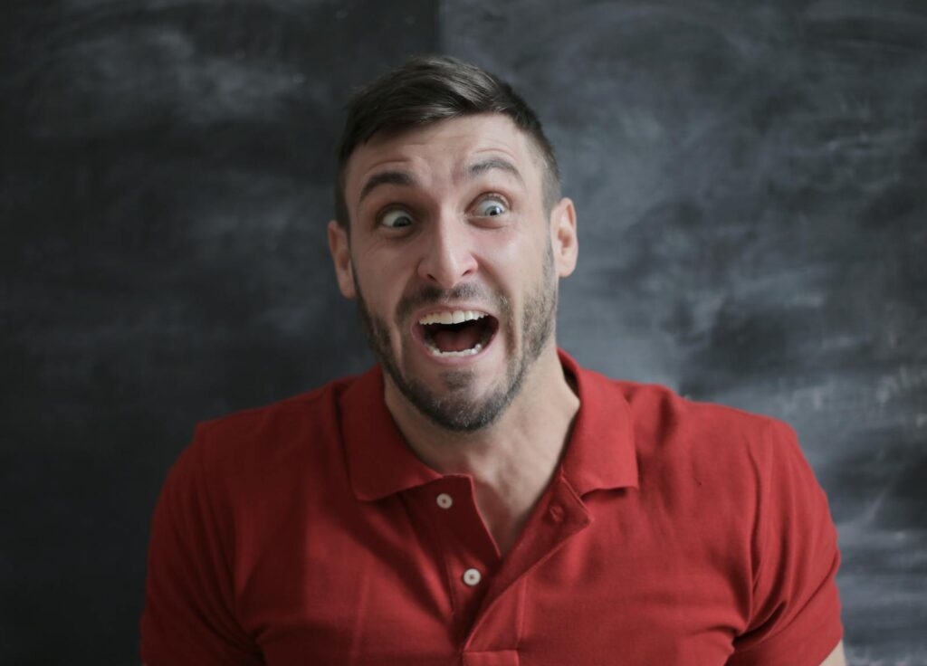 Expressive portrait of surprised man in red polo shirt with open mouth and wide eyes against a dark background.