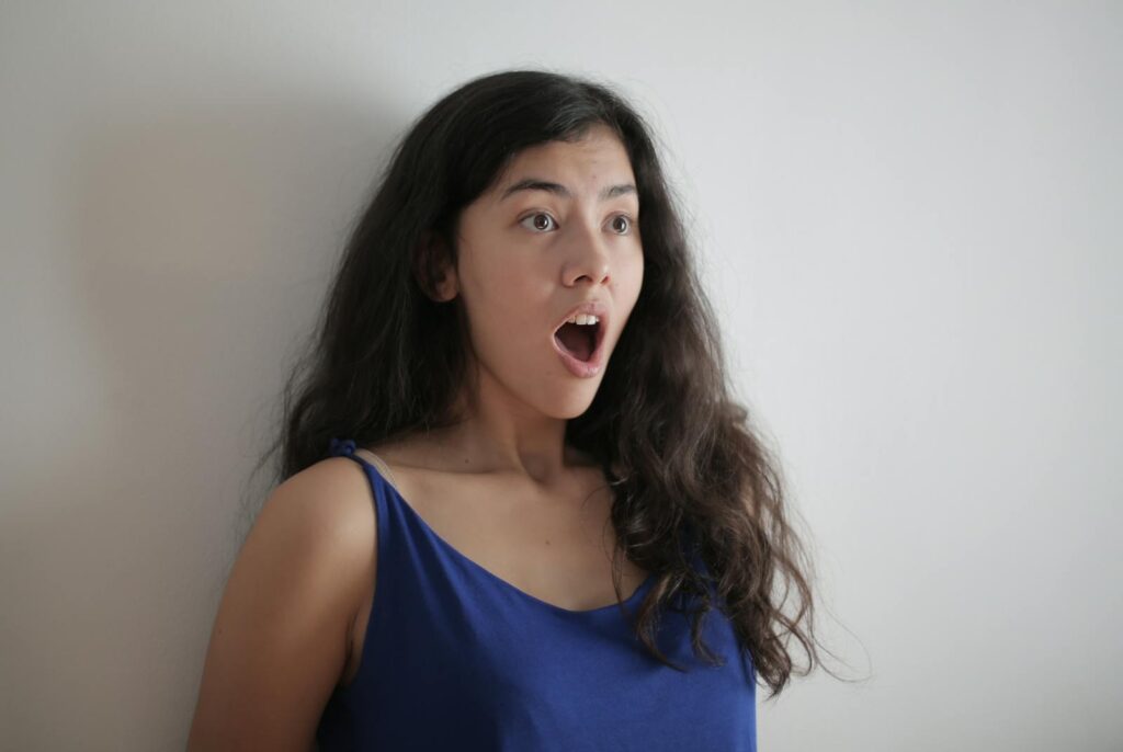 A surprised young woman with long brunette hair and an expressive face in a blue top.