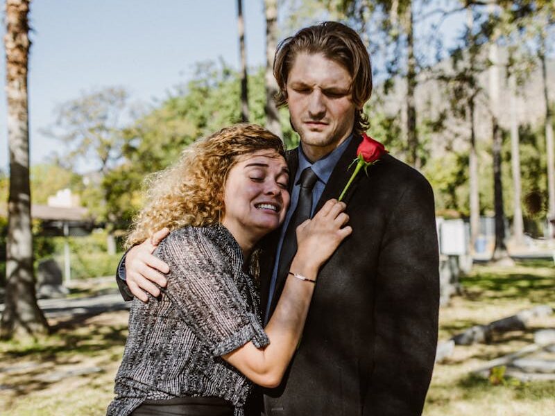 A couple in mourning, embracing in a cemetery setting on a sunny day.